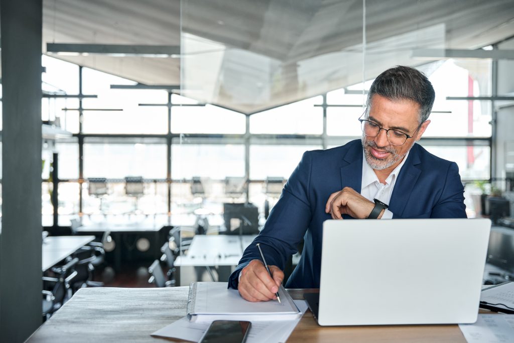 Middle aged smiling paralegal wearing suit sitting at desk in modern office working on Admin Tasks laptop computer and writing notes, copy space.